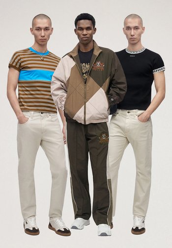 Trei bărbați stau unul lângă altul purtând ținute casual: tricouri cu dungi și negre cu pantaloni deschiși la culoare, și o jachetă bicoloră cu pantaloni asortați.