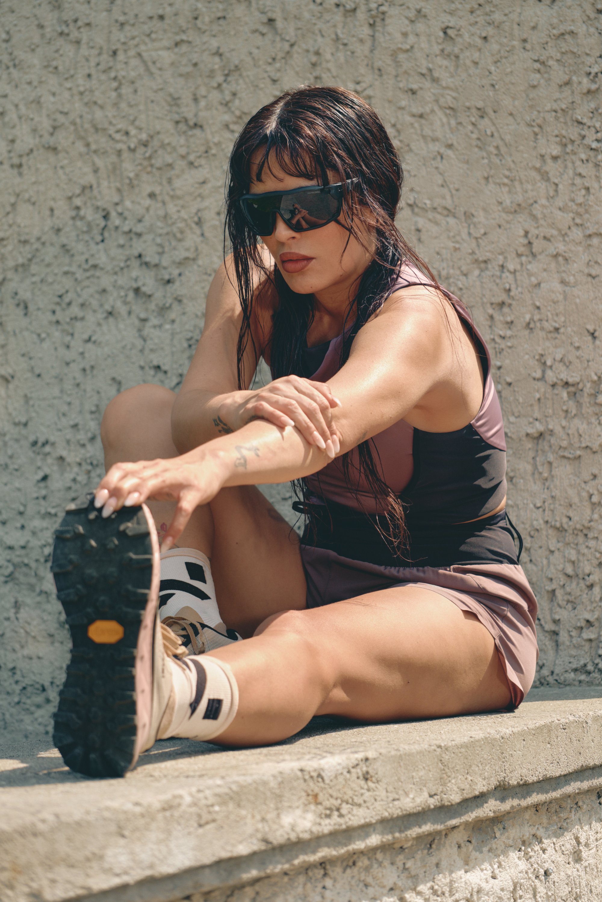 Conjunto atlético en negro y malva; lleva gafas de sol grandes y oscuras. Zapatillas de deporte color tostado con suela pronunciada y calcetines claros. Fondo de pared texturizada.