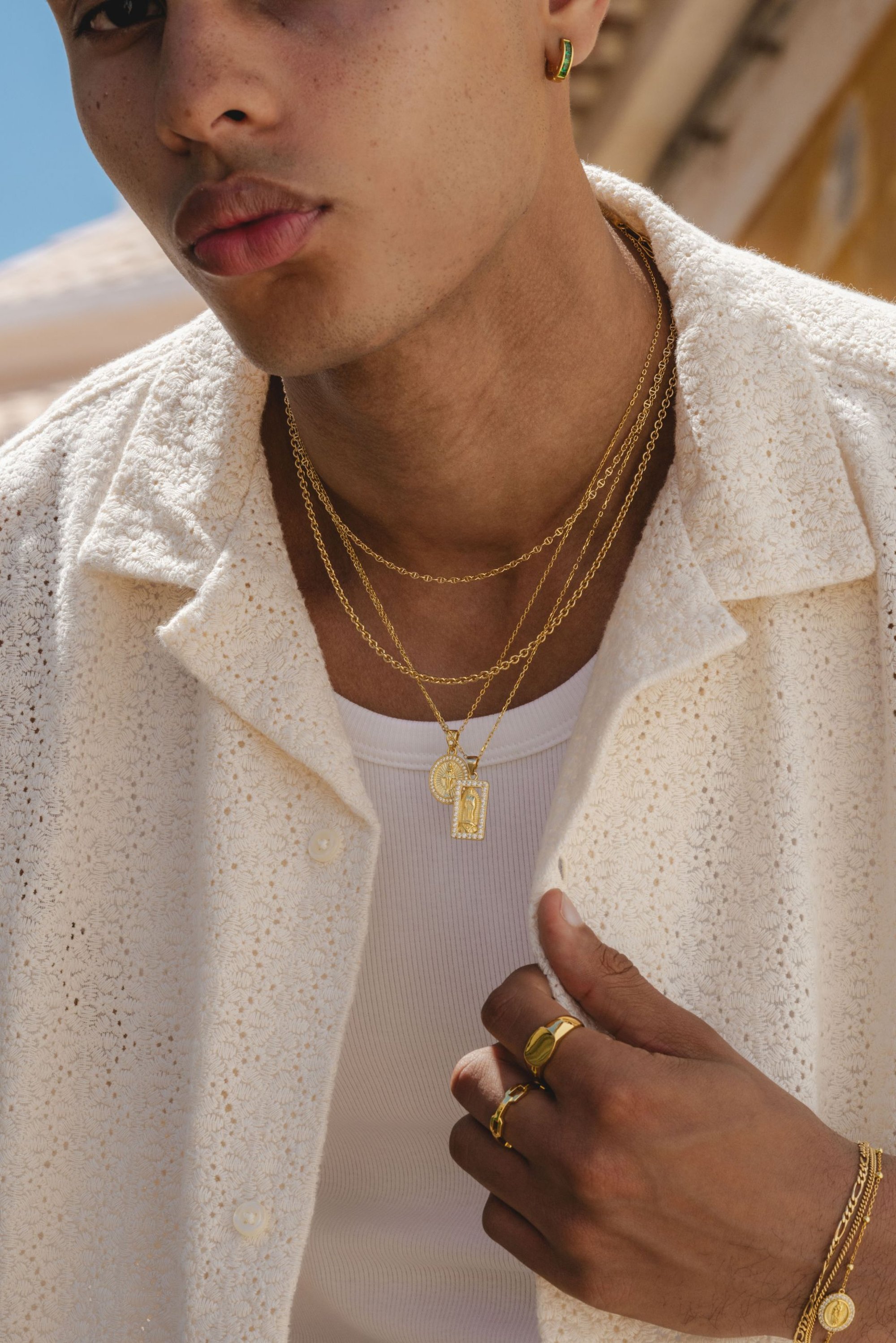 Camisa blanca texturizada de botones sobre una camiseta de tirantes blanca, con cadenas de oro en capas y múltiples anillos de oro en una mano bronceada.