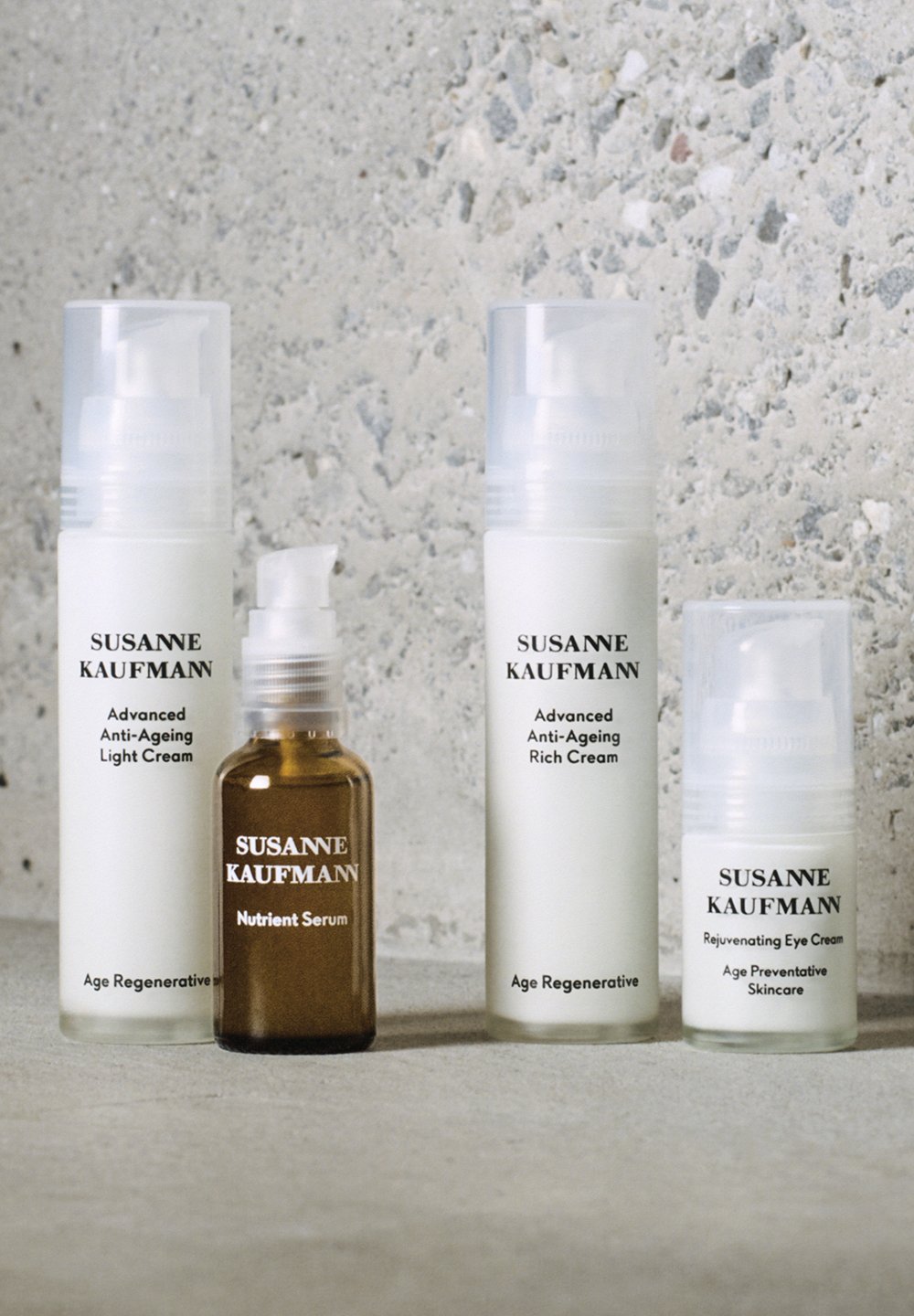 Quatre flacons de soins de la peau Susanne Kaufmann sur une surface grise : deux crèmes anti-âge, un sérum nutritif et une crème pour les yeux.