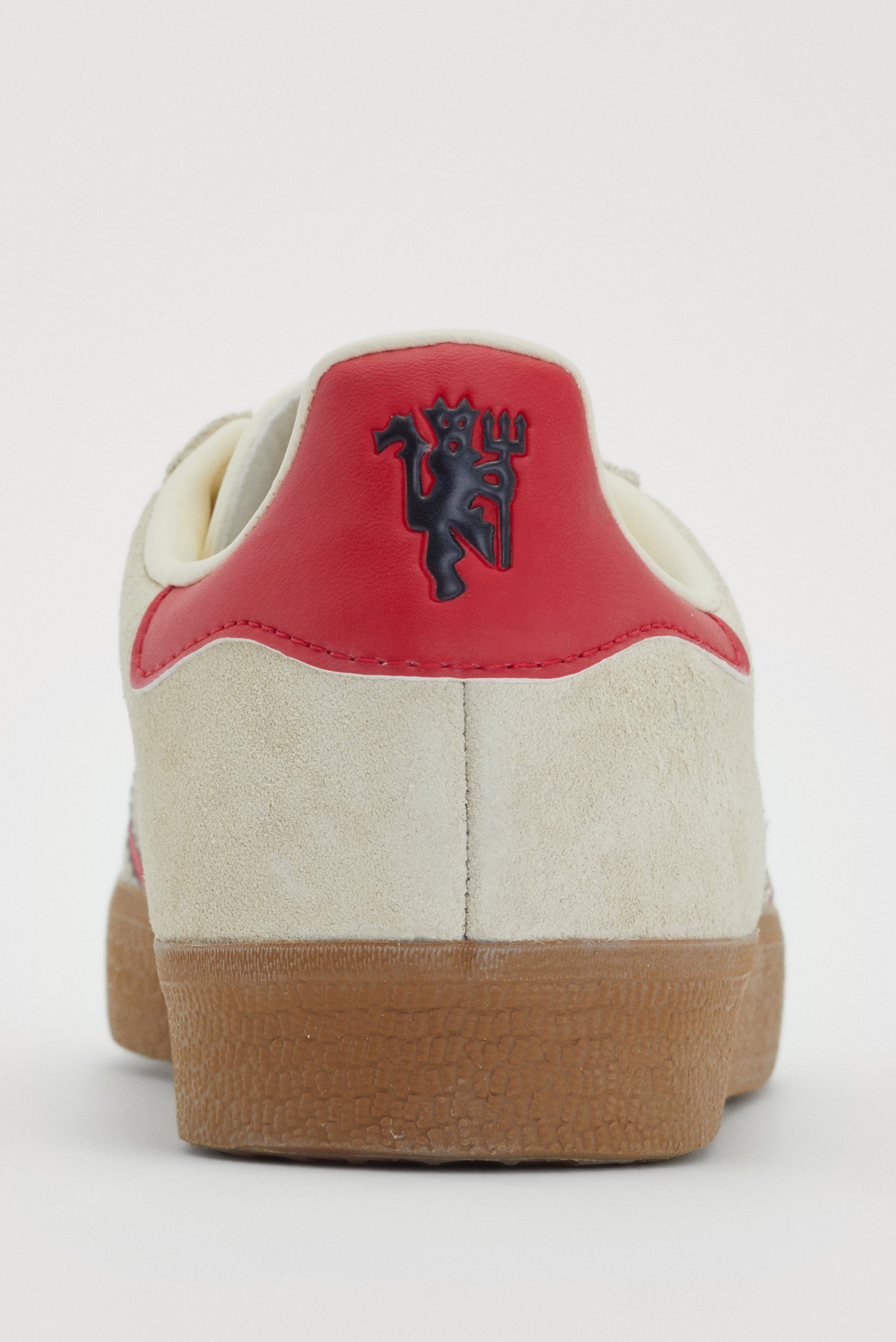 Achteraanzicht van een beige sneaker met rode accenten en een logo met een gestileerde figuur, met een getextureerde rubberen zool.
