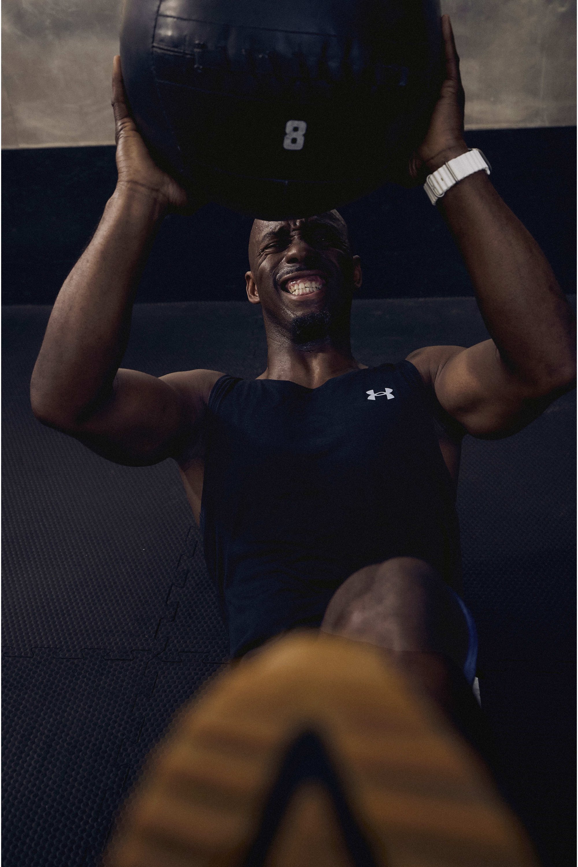 Svalnatý muž v čiernom športovom tričku zdvíha ťažkú medicinálnu loptu nad hlavu, usmievajúc sa pri námahe, v telocvični. Pod ním je viditeľná tmavá podlaha.