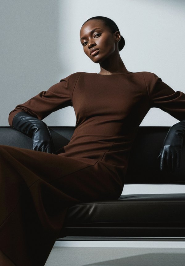 Brun figursydd klänning med långa ärmar, strukturerat tyg och rund halsringning. Modellen bär svarta läderhandskar och sitter på en svart soffa.