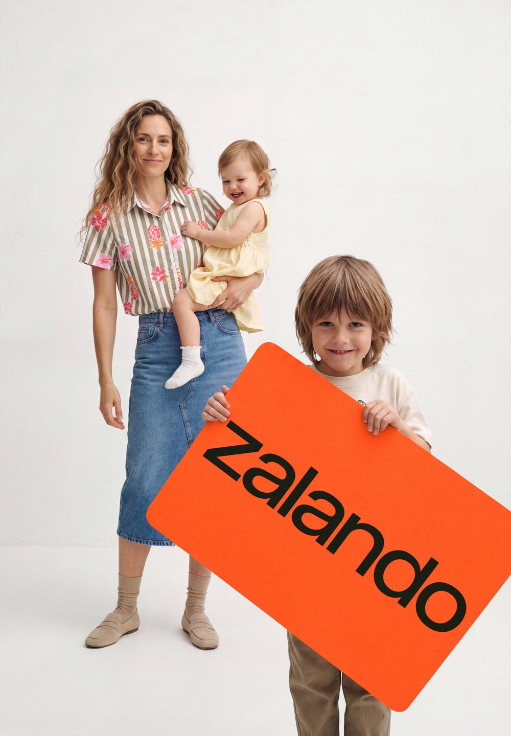 Lächelnde Frau mit Kleinkind steht hinter einem Jungen, der ein großes oranges Zalando-Schild vor einem einfarbigen weißen Hintergrund hält.