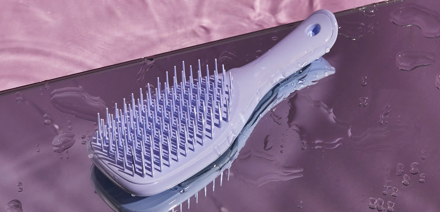 Lyse lilla plastik hårbørste med fleksible børster, der hviler på en reflekterende våd overflade med pink baggrund.
