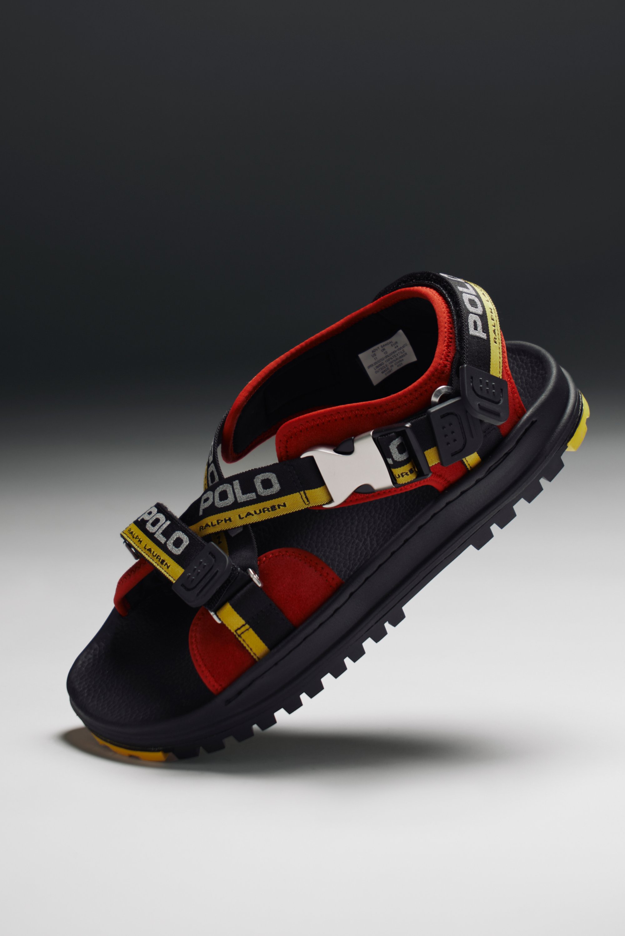Red and black sandal with a chunky sole and adjustable straps featuring a "Polo" logo in yellow. Suspended against a dark background.