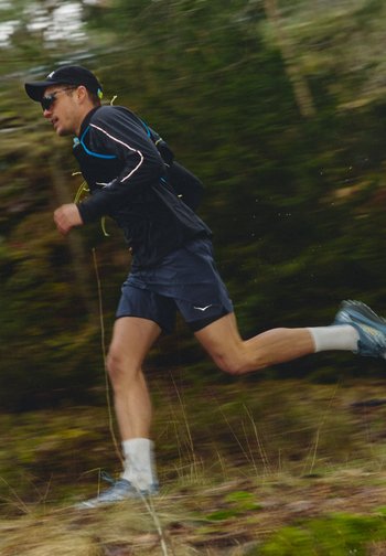 Mann med svart caps, solbriller og treningstøy som løper raskt gjennom en skogssti med uskarp bakgrunn.