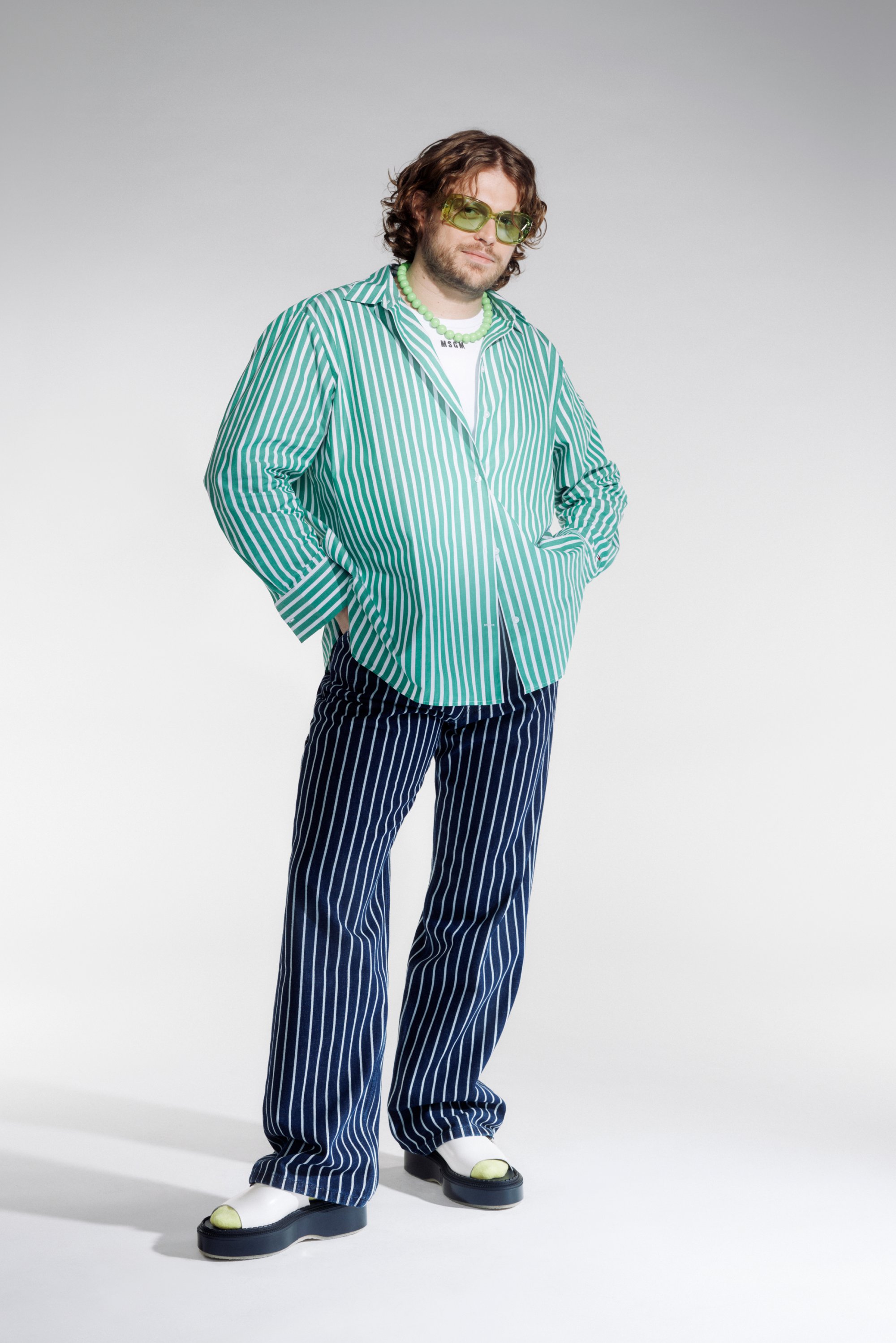 Een persoon met krullend haar draagt een groen-wit gestreepte shirt, marineblauwe gestreepte broek en groene zonnebril, terwijl hij met zijn handen in zijn zakken staat.