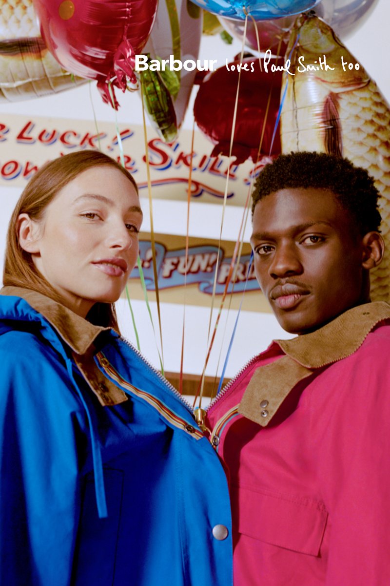 Dos modelos vestidos con chaquetas azul y roja con cuellos beige están juntos, sosteniendo globos de colores en un ambiente festivo de parque de diversiones.