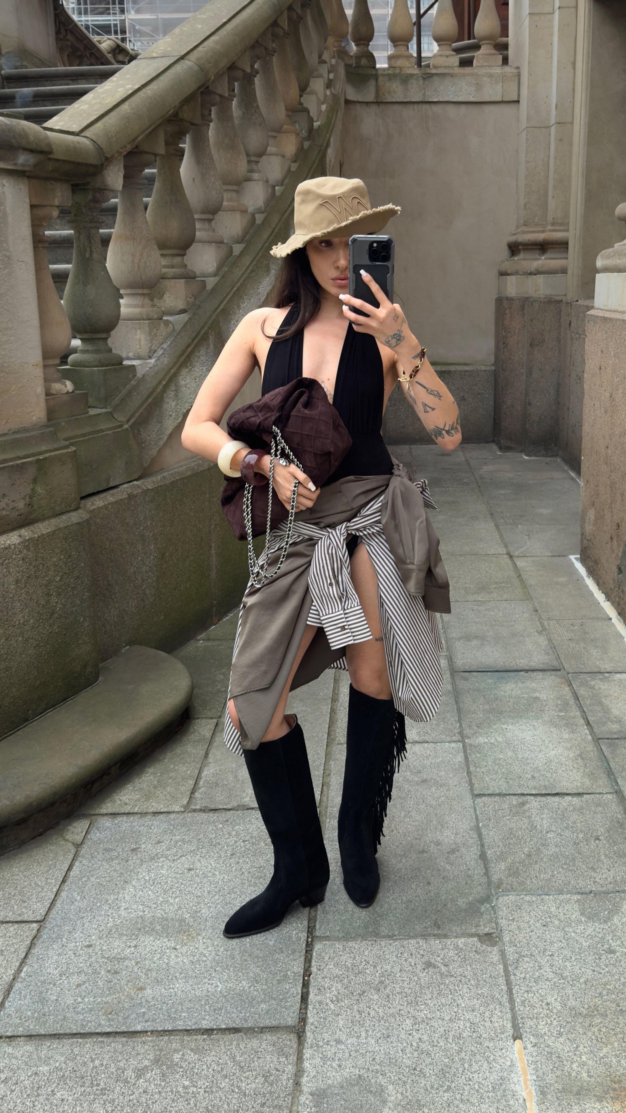 Costume da bagno nero, cappello a falda larga beige, abbigliamento a strati compreso un borsa marrone trapuntata e una camicia a righe grigie, e stivali neri in suede al ginocchio.