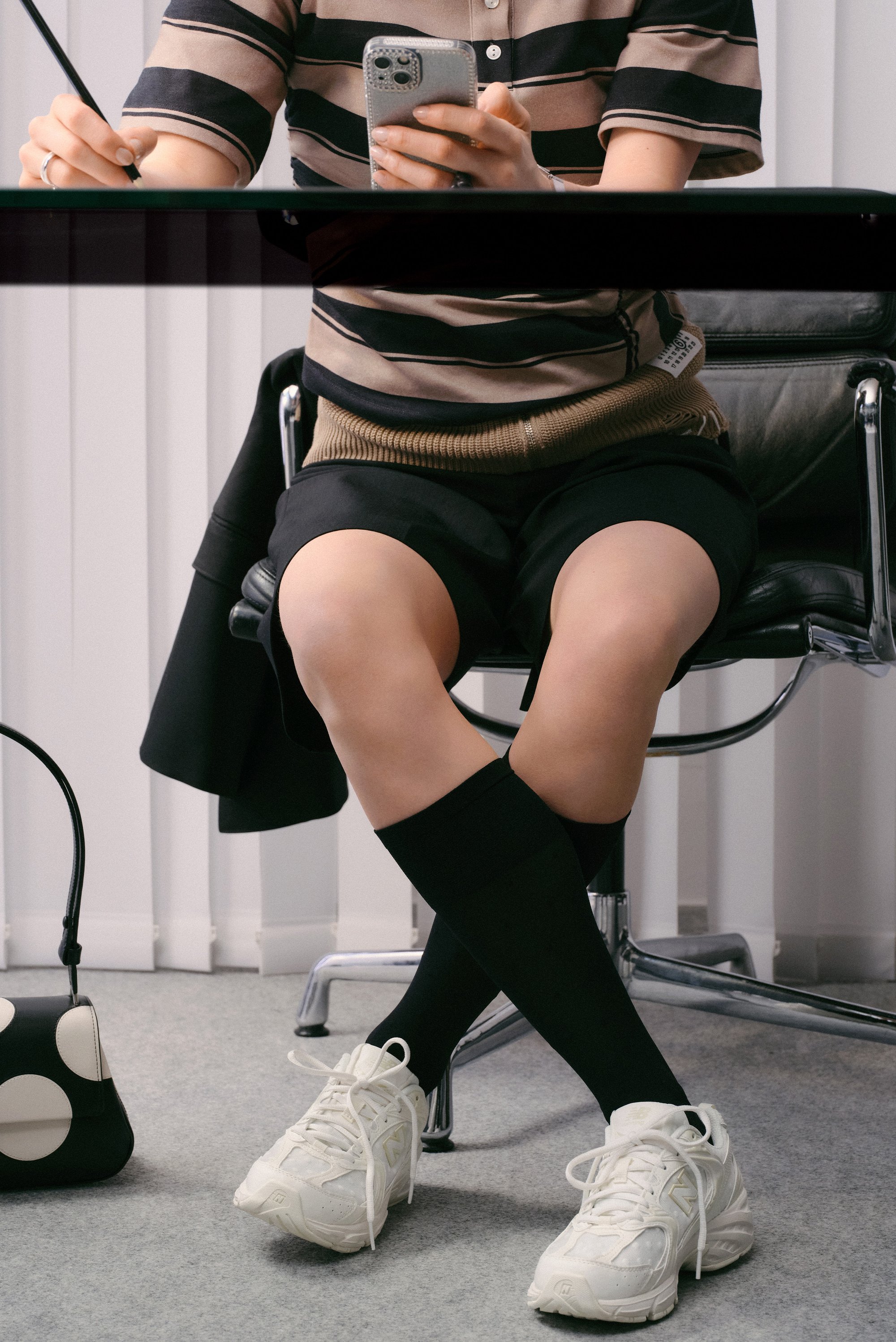 Una persona è seduta a una scrivania, indossando una polo a righe e pantaloni corti, mentre tiene uno smartphone. Ha calze fin sopra il ginocchio e sneakers bianche. Una borsa è appoggiata nelle vicinanze.