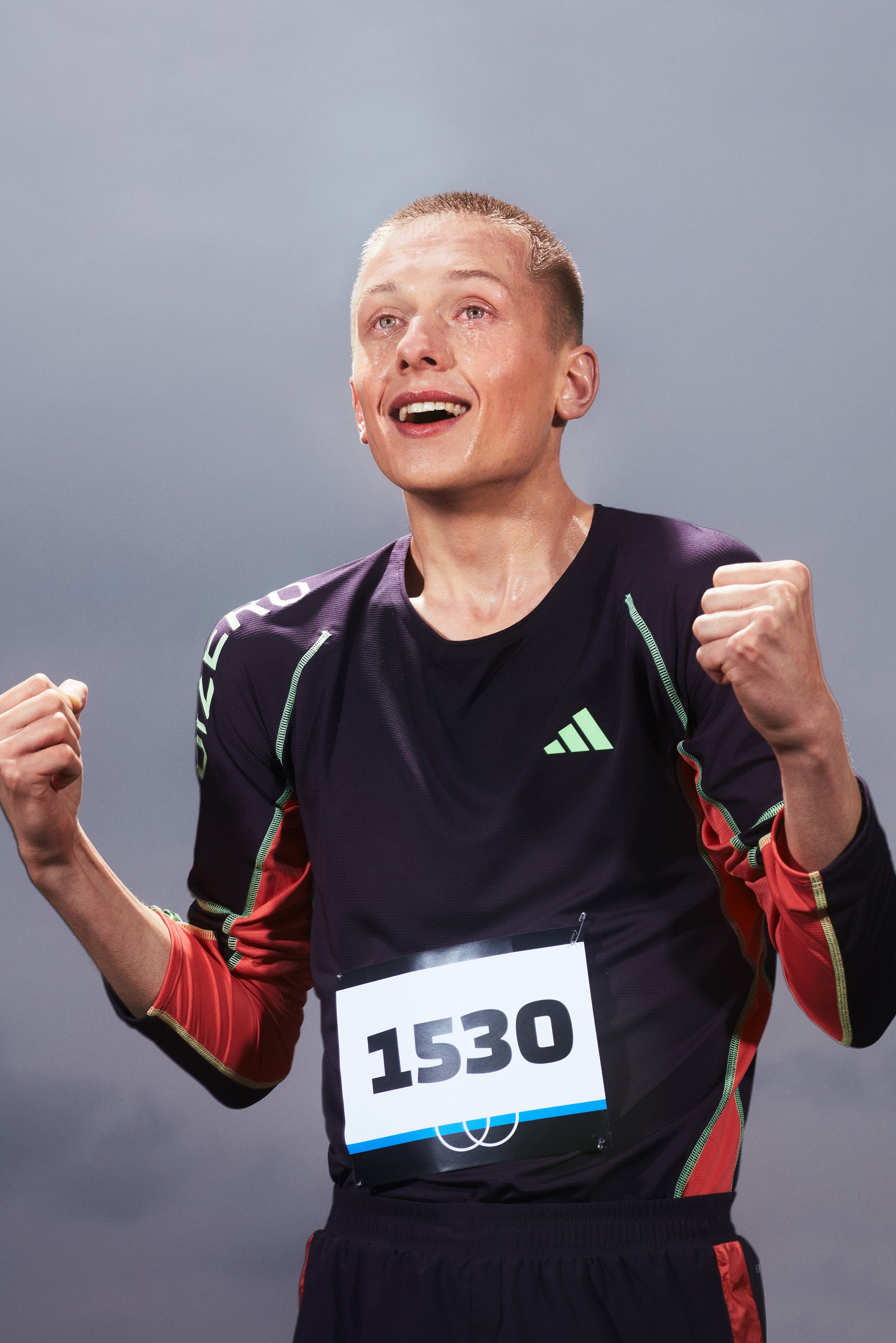 A young male athlete, wearing a black and red sports shirt and bib number 1530, celebrates with raised fists, beaming with joy.