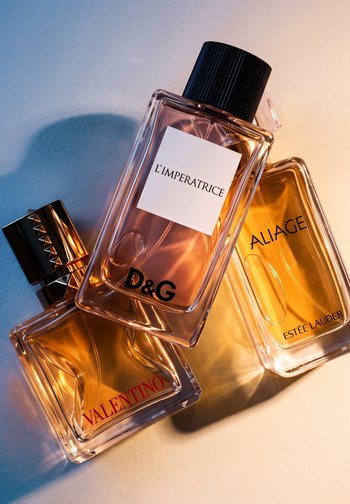Trzy prostokątne flakoniki perfum w bursztynowych i różowych tonach z etykietami Valentino, D&G L’Impératrice i Estée Lauder Aliage na jasnej powierzchni.