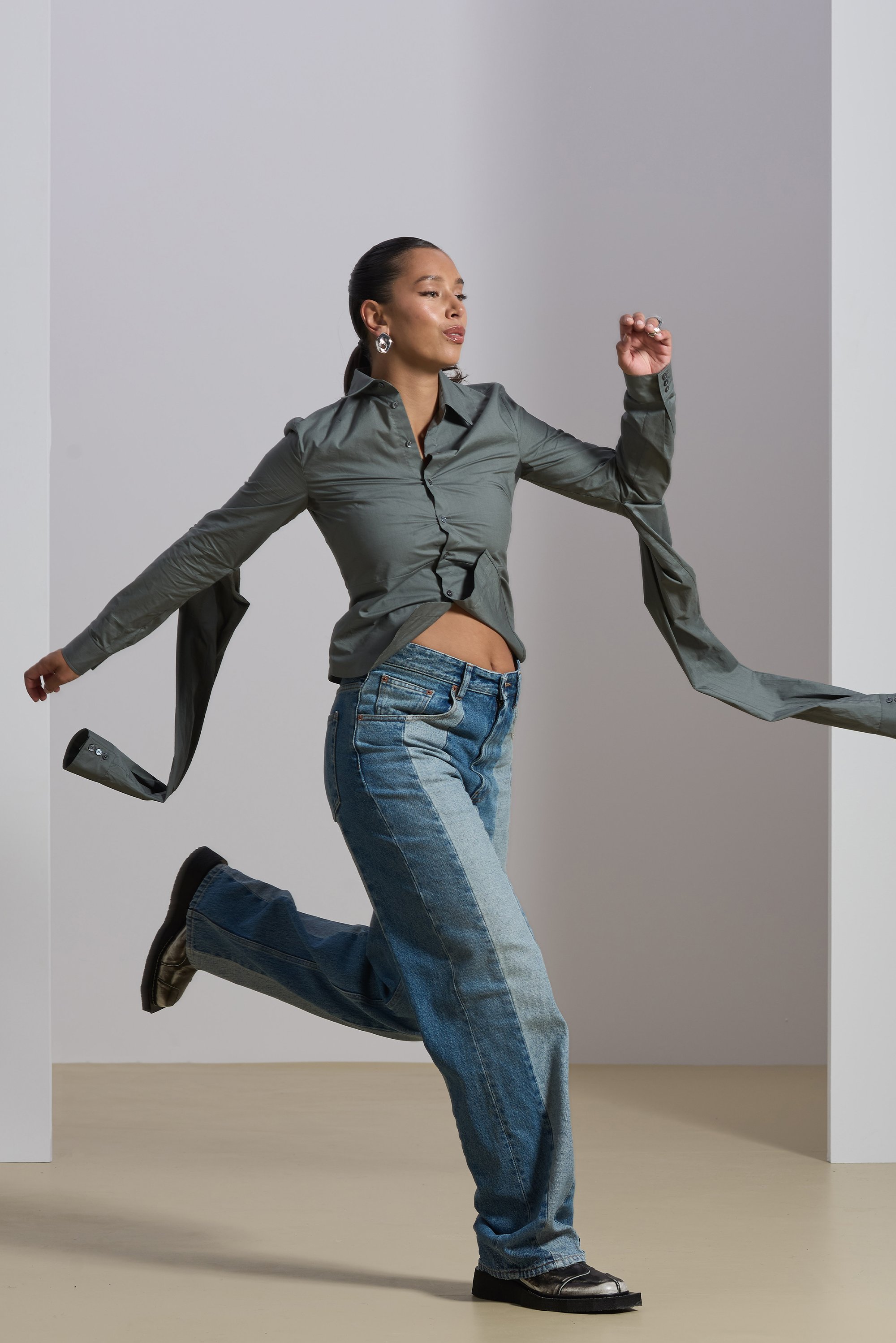 A model in a loose, light green shirt and wide denim jeans runs playfully, with flowing shirt sleeves, against a neutral background.