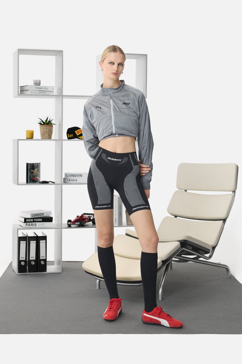 Chaqueta corta gris, pantalones cortos negros con detalles en blanco, calcetas negras hasta la rodilla y zapatillas rojas. Tejidos suaves, diseño atlético moderno.