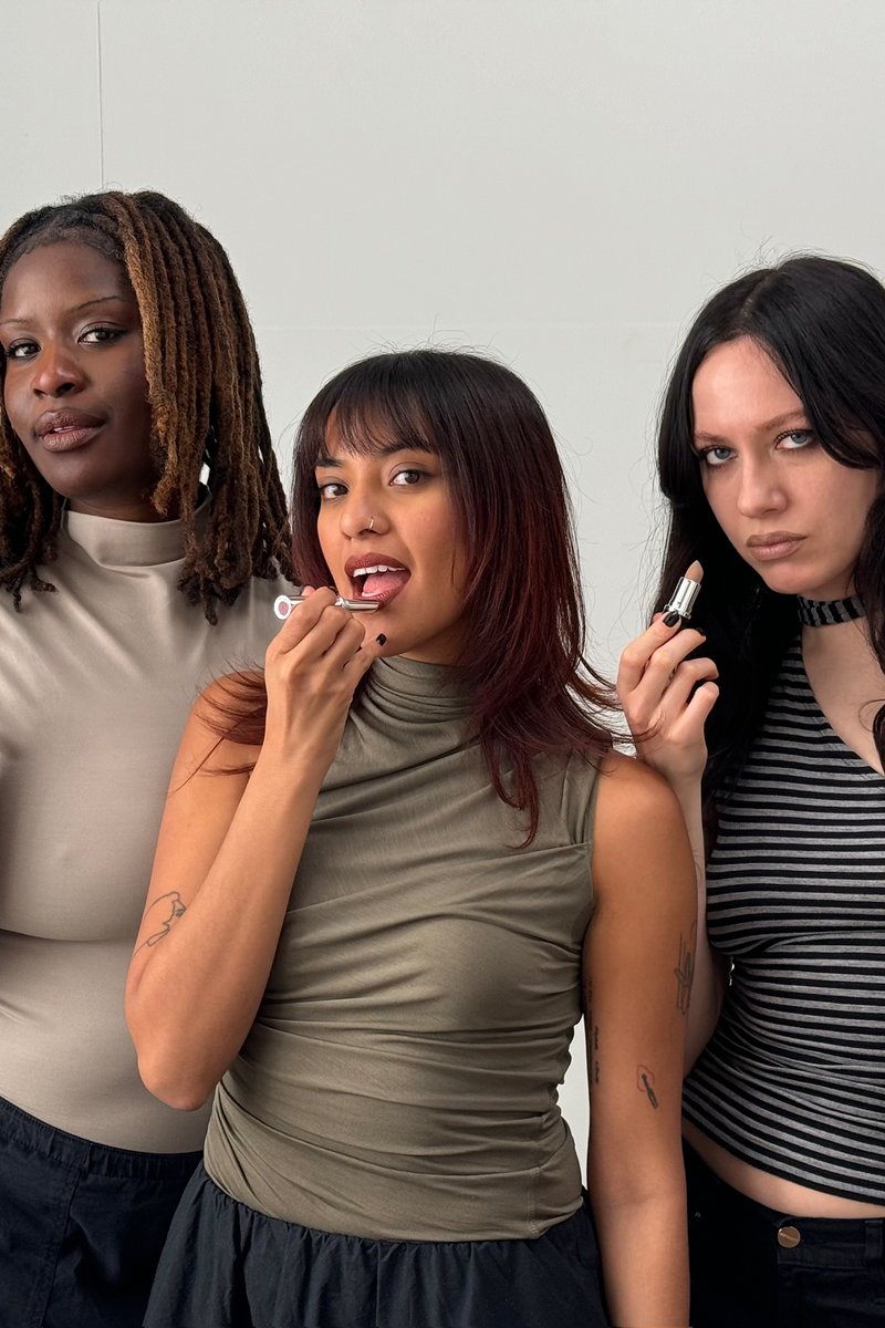 Three women pose together, applying lipstick. The woman in the center makes a playful face, while the others hold lipstick, showcasing varied styles.