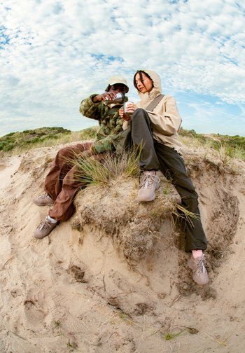 Twee personen zitten op een zanderige heuvel, waarbij de een een drankje in een kopje giet dat door de ander wordt vastgehouden, omgeven door gras en een bewolkte lucht.