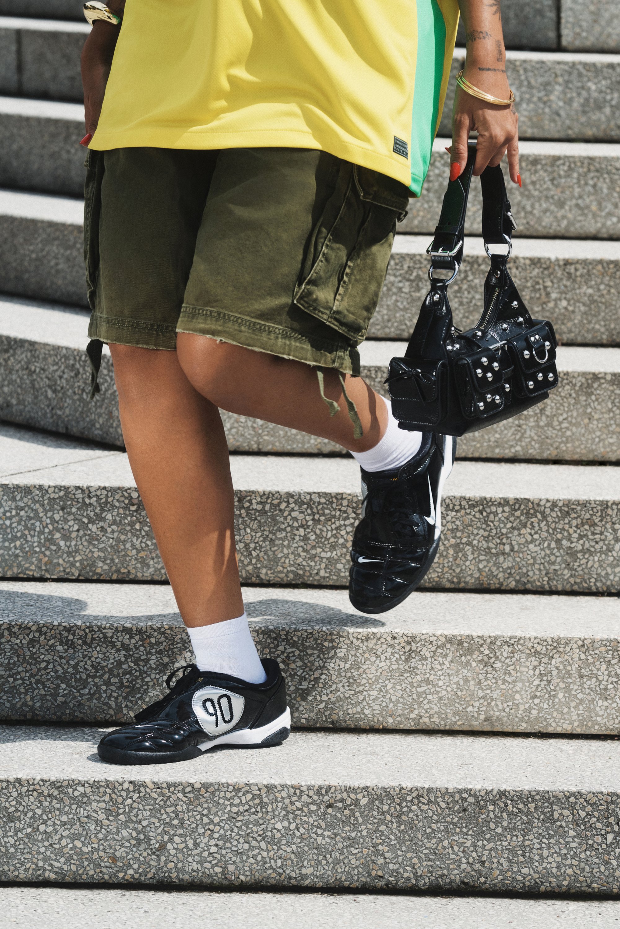Zapatillas deportivas negras con acentos blancos y el número "90," combinadas con pantalones cortos tipo cargo en color verde oliva y un bolso negro con tachuelas.