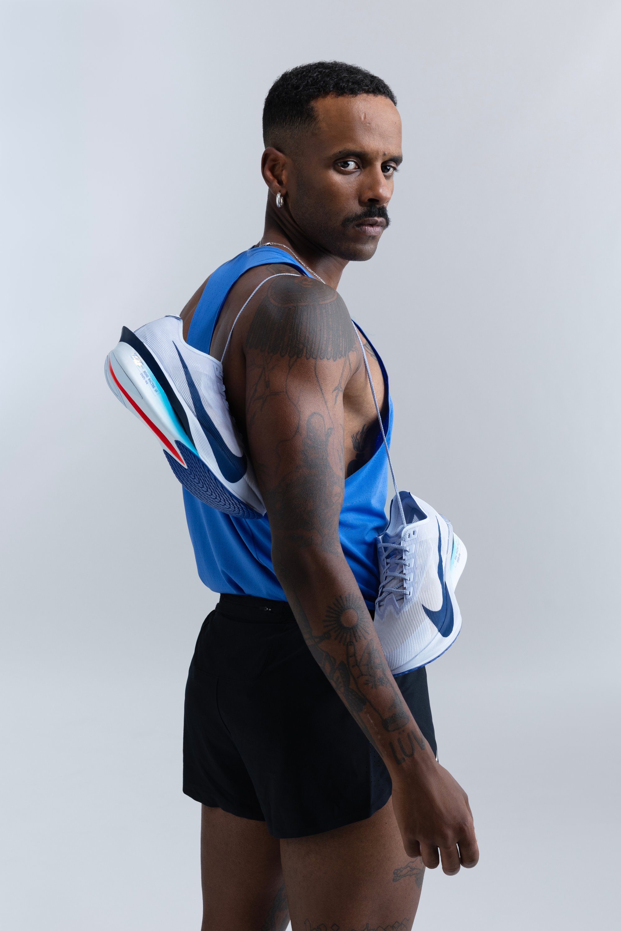 Man met tatoeages draagt een blauwe tanktop en zwarte shorts, en draagt witte hardloopschoenen met een blauwe swoosh over zijn schouder tegen een effen achtergrond.