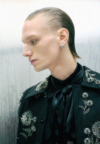 Black embellished jacket featuring floral silver details, worn over a black silk blouse with a necktie and a silver earring shaped like a flower.