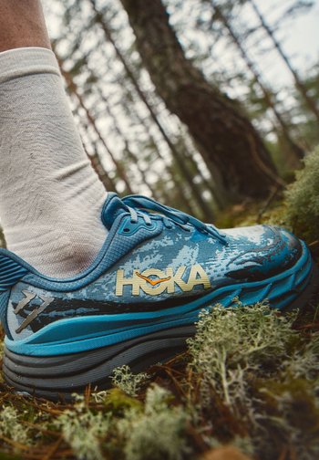 Blå Hoka løpesko med gjørme på, brukt med hvit sokk, stående på mosegrodd skogbunn med trær i bakgrunnen.