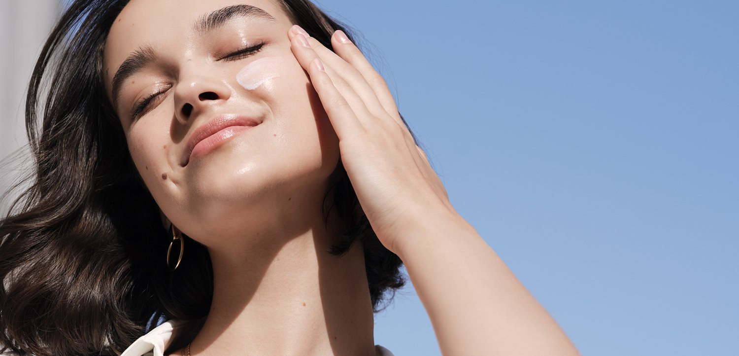 Woman with closed eyes applies cream to her cheek against a clear blue sky background, wearing gold hoop earrings.