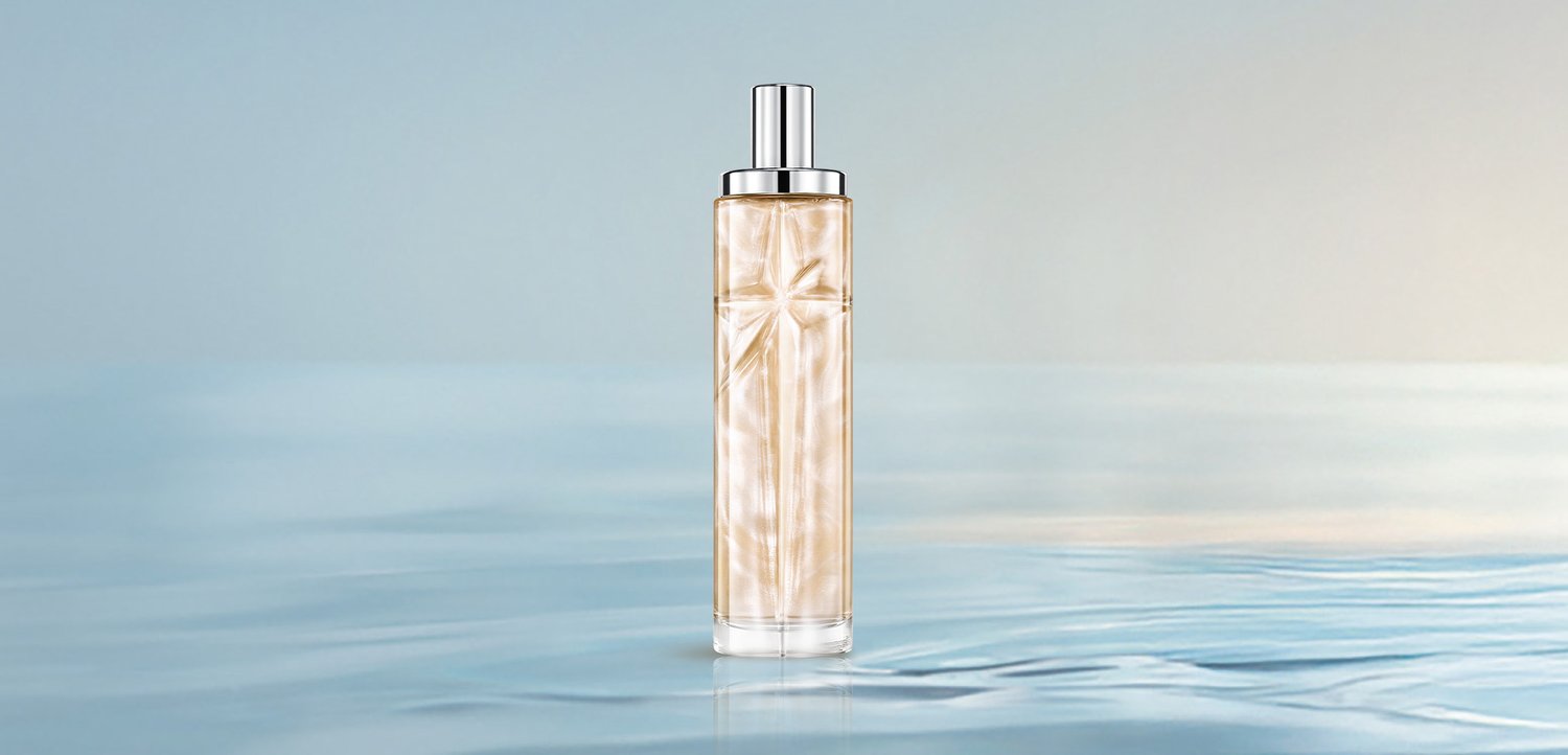 Botella de perfume alta y transparente con líquido dorado y tapa plateada, situada sobre una superficie de agua tranquila con un fondo suave azul y beige.