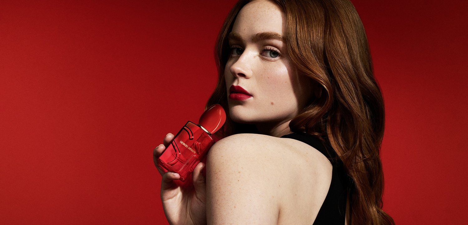 Red perfume bottle with a rounded cap, featuring a sleek design and embossed label. Model with long, wavy reddish-brown hair poses against a red background.