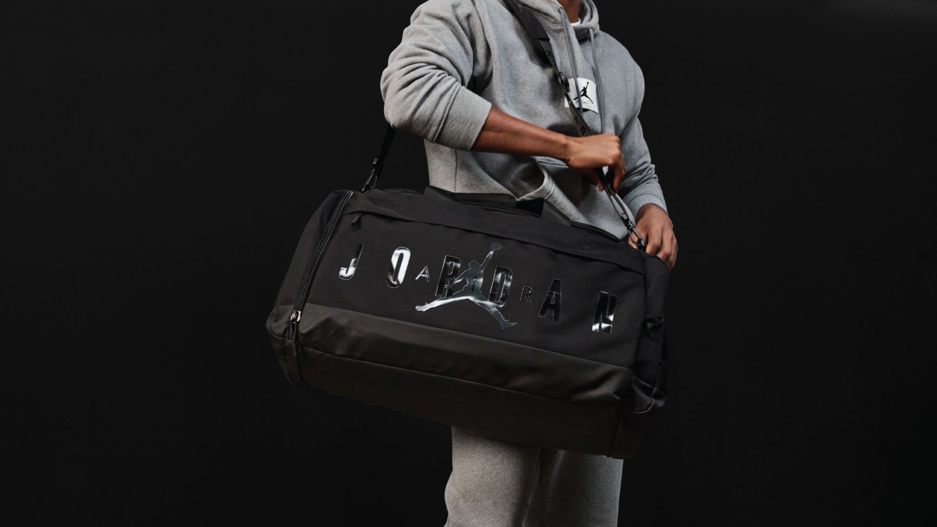 Une personne portant un sweat à capuche gris et un pantalon de survêtement tient un sac de sport noir avec un logo Jordan réfléchissant sur fond noir.
