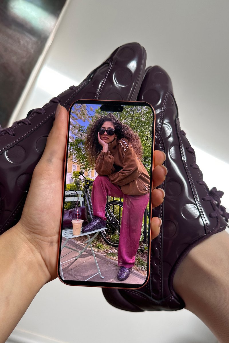 Hand hält Telefon, auf dem eine Frau mit lockigem Haar, Sonnenbrille, brauner Jacke, rosa Hose und dunkelvioletten Schuhen zu sehen ist, die ihren Fuß auf den Tisch mit Getränk legt.