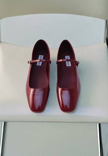 Glanzende rode lakleren Mary Jane schoenen met een vierkante neus en een dunne band met een kleine gesp, tentoongesteld op een witte zetel.