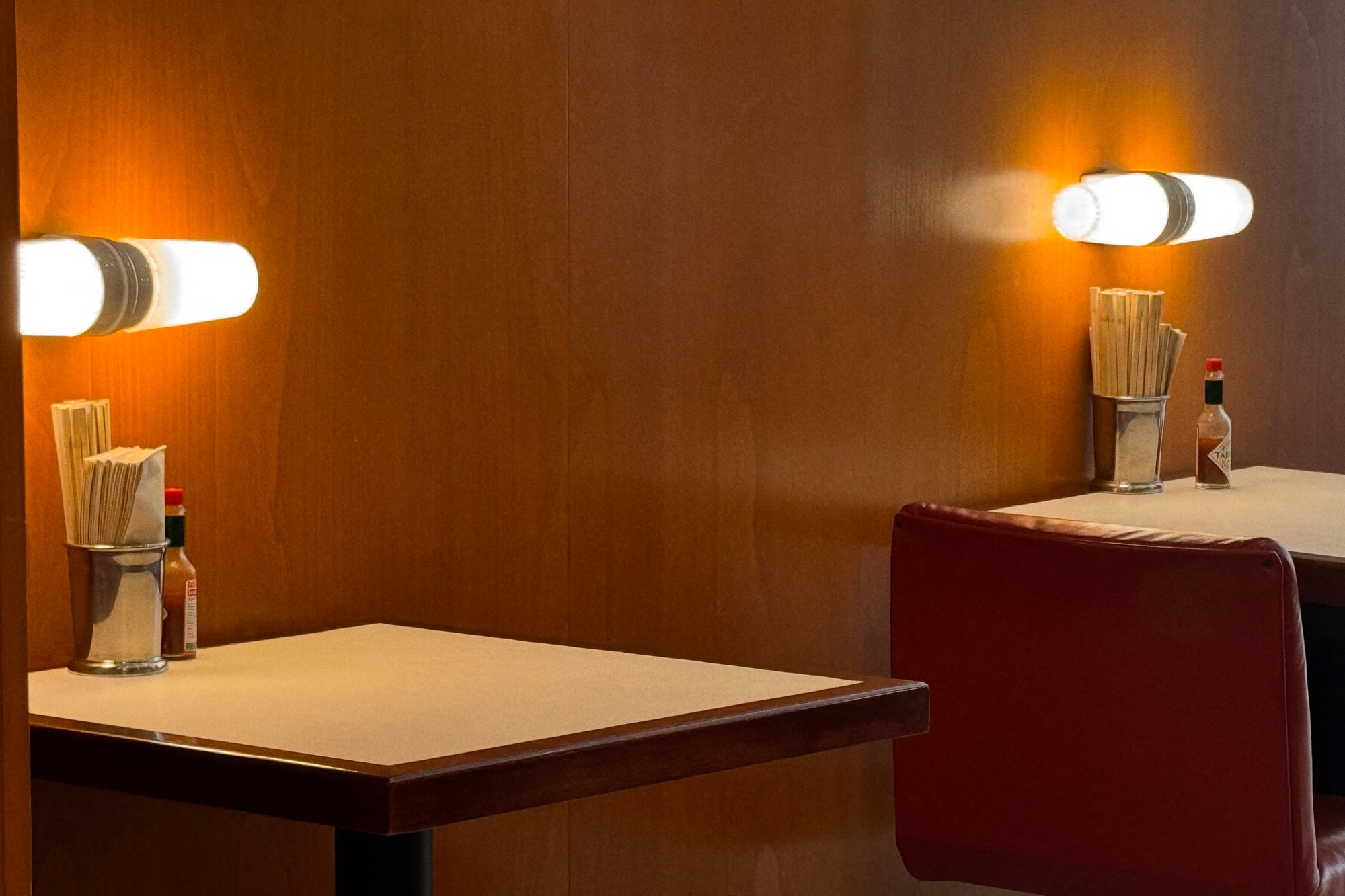 Dos mesas en un ambiente acogedor de restaurante presentan condimentos y utensilios, iluminados por luces montadas en la pared contra paneles de madera.