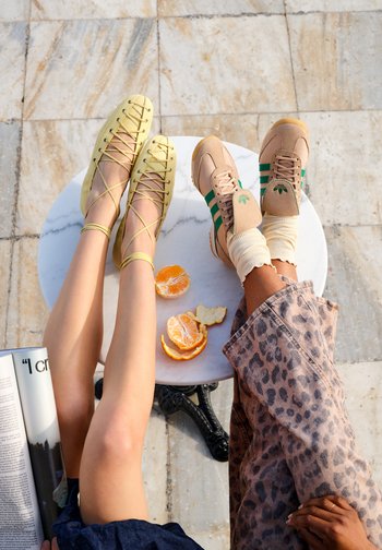 Dva páry nohou odpočívající na stole s oloupaným pomerančem; jedny mají žluté baleríny se šněrováním, druhé růžové tenisky se zelenými pruhy a ponožkami.