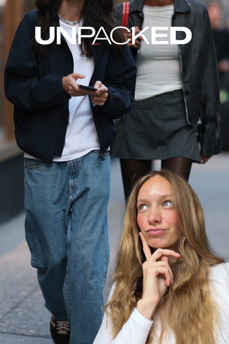 Mujer rubia de cabello largo y camisa blanca se toca el mentón pensativamente, con dos personas vestidas informalmente caminando detrás de ella en una calle de la ciudad.