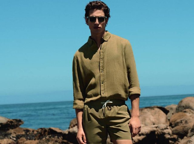 Homme portant des lunettes de soleil, une chemise olive boutonnée et un short, debout sur une rive rocheuse avec l'océan et un ciel dégagé en arrière-plan.