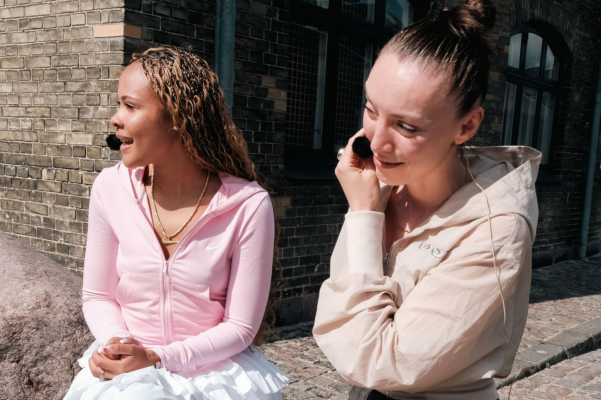 Twee personen zitten tegen een bakstenen muur. De een draagt een lichtroze hoodie met rits en een voorzak, de ander in een beige hoodie. Beide hebben gekruld haar.