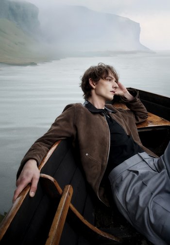 Veste en daim marron avec un col noir, portée sur une chemise noire. Pantalons plissés gris, assis dans un bateau en bois sur un lac brumeux avec des falaises en arrière-plan.