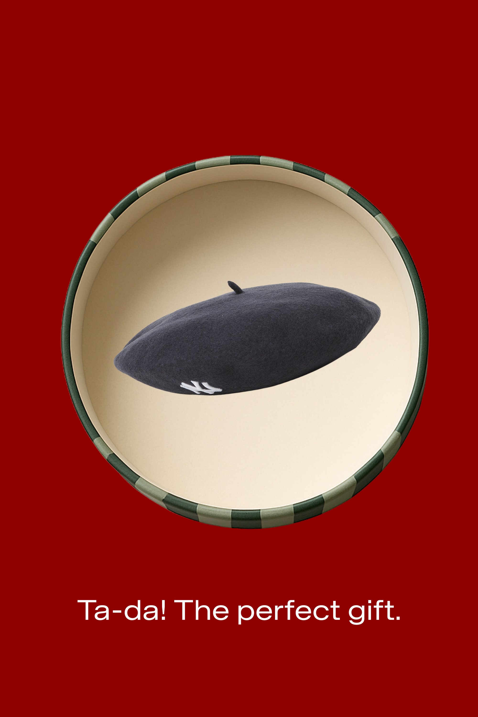 Dark grey beret with white logo inside a round gift box with a green striped rim on a red background, text below reads "Ta-da! The perfect gift."