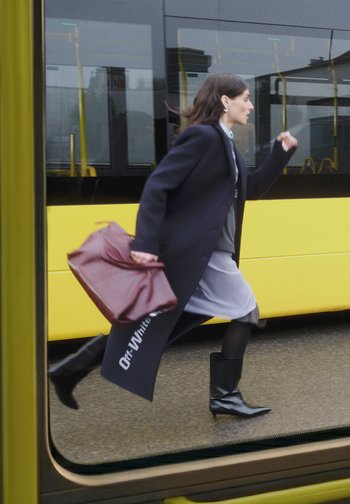 Bordeauxrode leren tas, zwarte kniehoge laarzen, lange marineblauwe jas, grijze jurk, rennend figuur voor een geel tramraam.