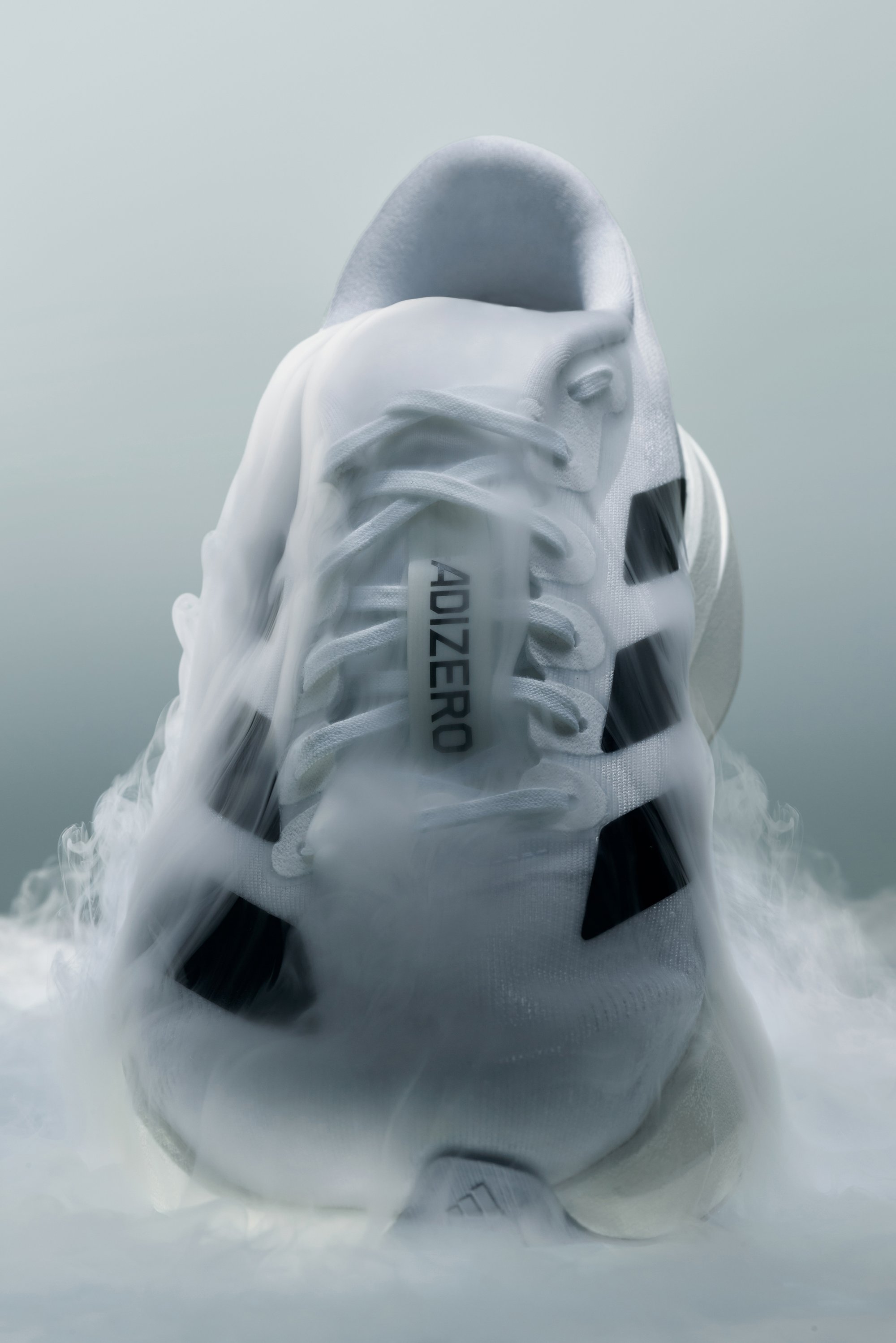 Zapatilla deportiva blanca parcialmente envuelta en niebla, con detalles en negro y el logo "ADIZERO" que se muestra de manera prominente en el lado.