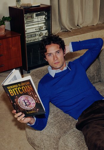 Modell i en blå genser med skjorte, holder en bok med tittelen "Bitcoin" med et rundt logo på omslaget, sittende på en teksturert sofa.