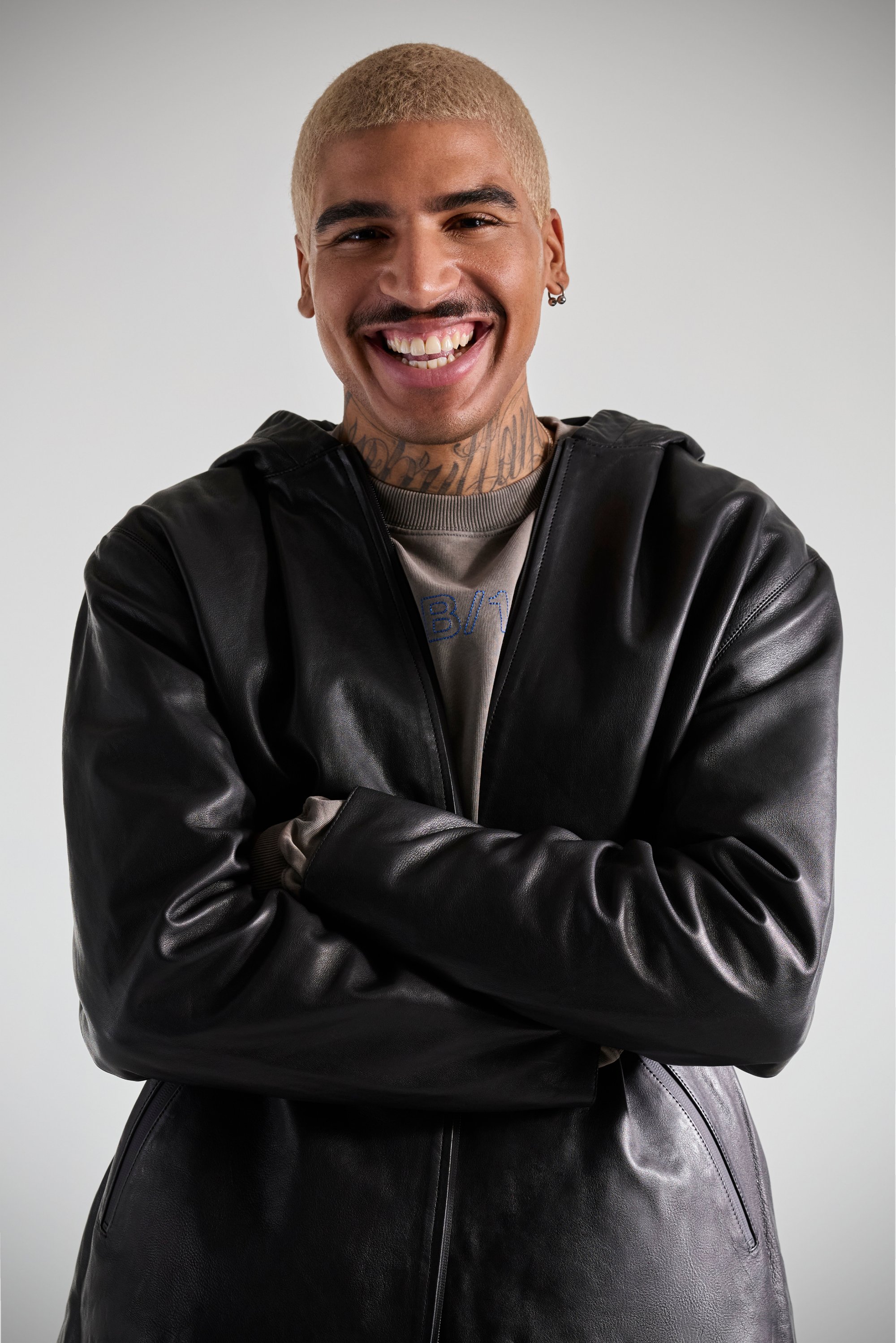 Person with short blonde hair and tattoos smiles, wearing a black leather jacket over a light-colored shirt. Background is neutral.