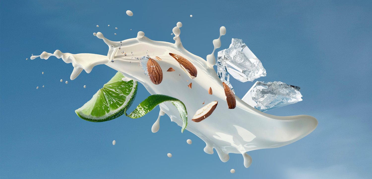Spetterende melk met amandelen, limoenpartjes en ijsblokjes tegen een blauwe achtergrond, wat een dynamische en frisse compositie creëert.