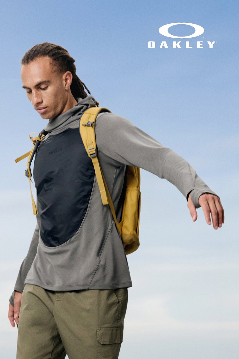Person with long hair wears a grey athletic top and yellow backpack, posing against a blue sky background. Oakley logo visible.
