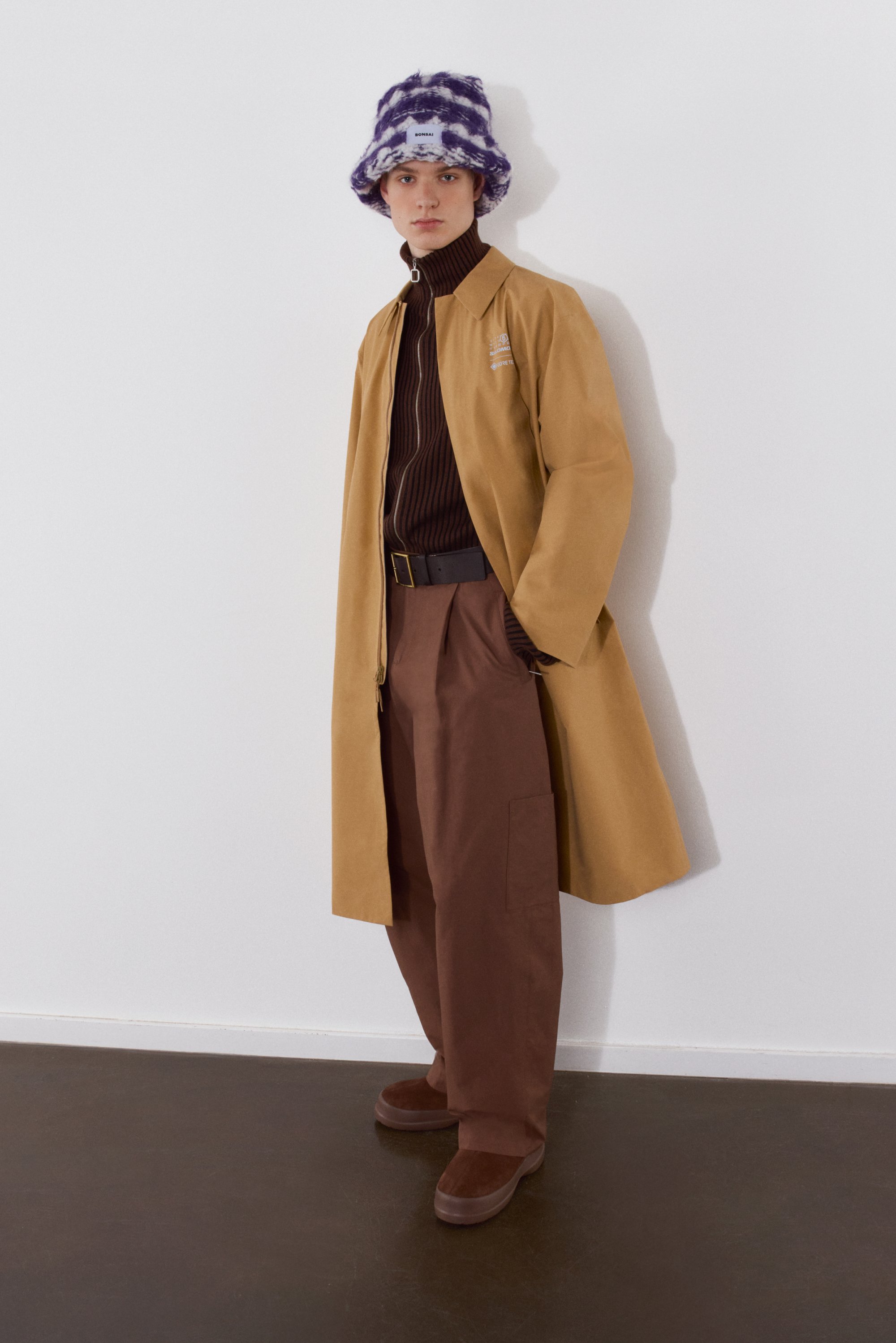 Una joven modelo está de pie contra una pared blanca, vistiendo un suéter de cuello alto marrón, un abrigo beige, pantalones de pierna ancha marrones y un sombrero de cubo morado con estampados.
