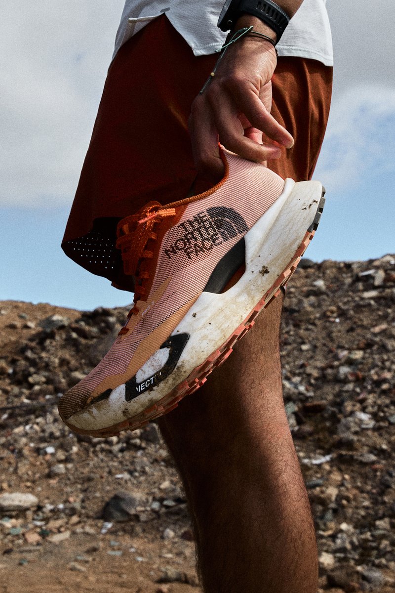 Person hält einen orangefarbenen The North Face Laufschuh mit schmutziger Sohle und steht auf felsigem Außengelände.