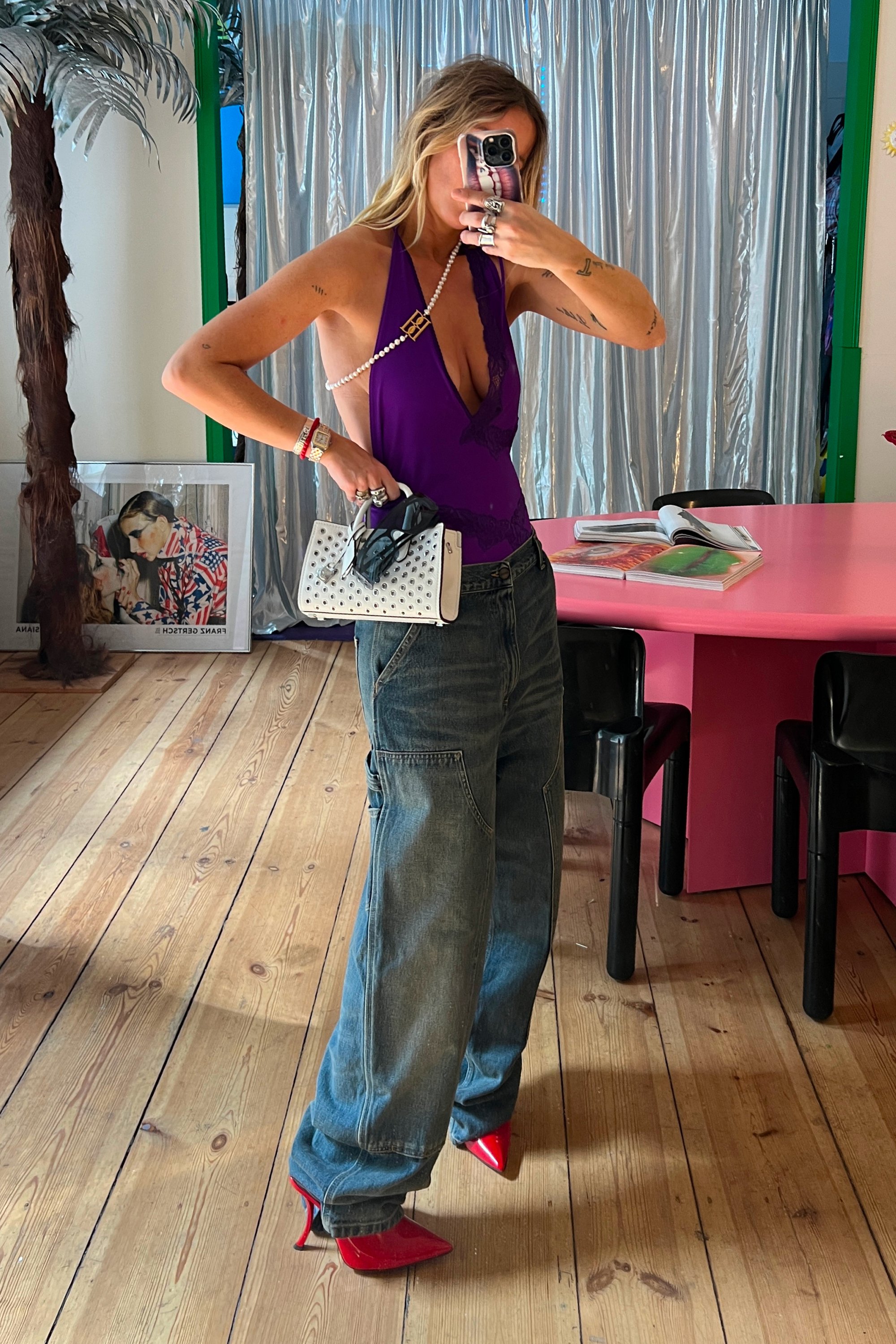 Mujer con un top halter púrpura y jeans anchos tomando un selfie en el espejo, sosteniendo un bolso blanco con gafas de sol, de pie sobre un suelo de madera cerca de una mesa rosa.