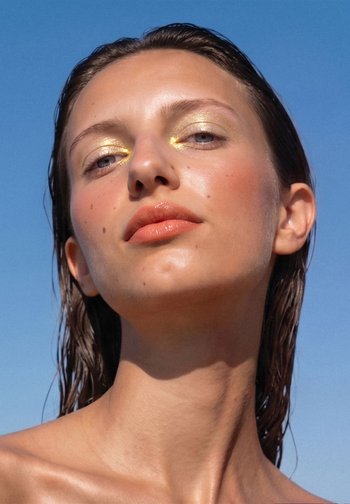 Femme aux cheveux mouillés et au maquillage doré des yeux regarde la caméra sur fond de ciel bleu clair.