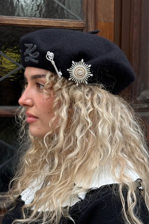 Sort filt beret med sølvdetaljer, herunder et solstrålebroche og en dekorativ palle. Afslappet krøllet hår delvist synligt.