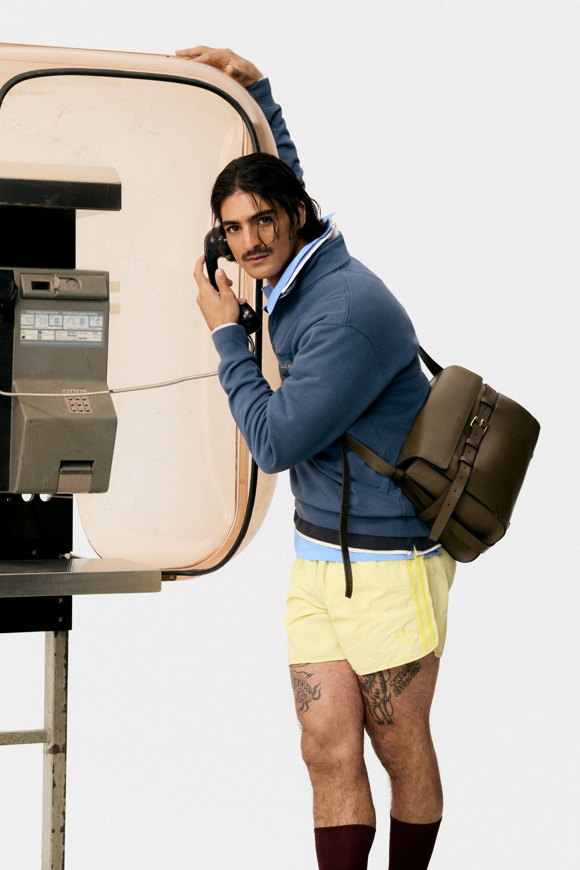 Een man met een snor houdt een vintage telefoon vast in een telefooncel, gekleed in een blauwe sweatshirt en gele shorts, met een rugzak over één schouder.